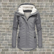 Giacca invernale da donna grigia con cappuccio foderato in pelliccia sintetica, chiusura a zip, tasche frontali, su sfondo di mattoni grigi.