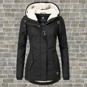Giacca invernale nera da donna con cappuccio foderato in pelliccia sintetica, chiusura a zip, tasche frontali, su sfondo di mattoni grigi.