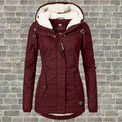 Giacca invernale da donna bordeaux con cappuccio foderato in pelliccia sintetica, chiusura a zip, bottoni decorativi, su sfondo di mattoni grigi.