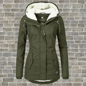 Giacca invernale verde oliva da donna con cappuccio foderato in pelliccia sintetica, chiusura a zip e bottoni, su sfondo di mattoni grigi.