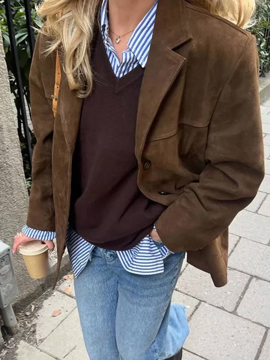 Donna con giacca marrone, camicia a righe blu, maglione marrone, jeans e caffè in mano. Moda autunnale casual chic.