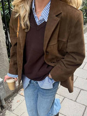 Donna con giacca marrone, camicia a righe blu, maglione marrone, jeans e caffè in mano. Moda autunnale casual chic.