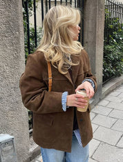 Donna bionda con giacca marrone in pelle scamosciata, camicia a righe blu, jeans, tiene caffè in mano. Moda autunnale casual in città.