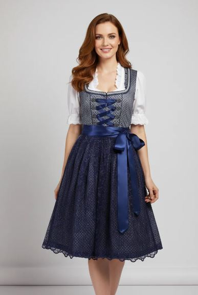 Donna sorridente in abito tradizionale dirndl blu con corpetto ricamato e fiocco, camicia bianca a maniche corte, sfondo grigio chiaro.