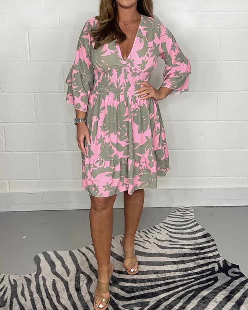 Donna con abito floreale rosa e verde, maniche a tre quarti, scollo a V, su sfondo bianco. Scarpe beige, tappeto zebrato. Moda femminile elegante.