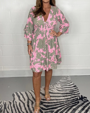 Donna con abito floreale rosa e verde, maniche a tre quarti, scollo a V, su sfondo bianco. Scarpe beige, tappeto zebrato. Moda femminile elegante.