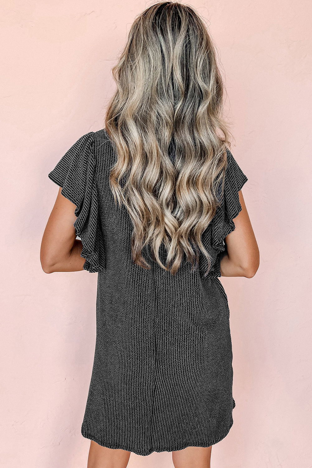 Donna con capelli ondulati biondi indossa un abito nero a coste con maniche corte a volant, sfondo rosa. Moda femminile, abbigliamento estivo elegante.