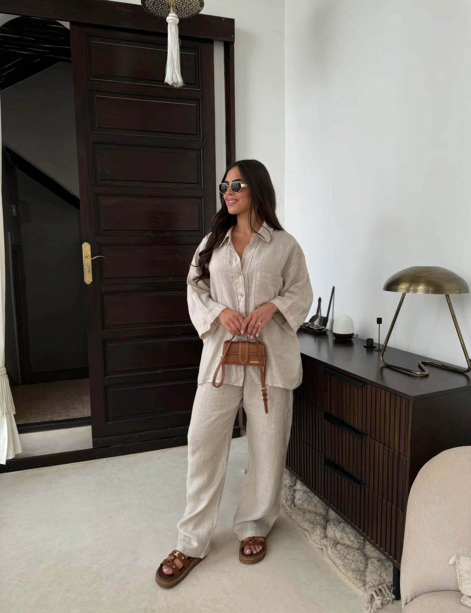 Donna con occhiali da sole indossa camicia e pantaloni beige, sandali marroni, borsa a tracolla. Sfondo interno moderno con porta e lampada.