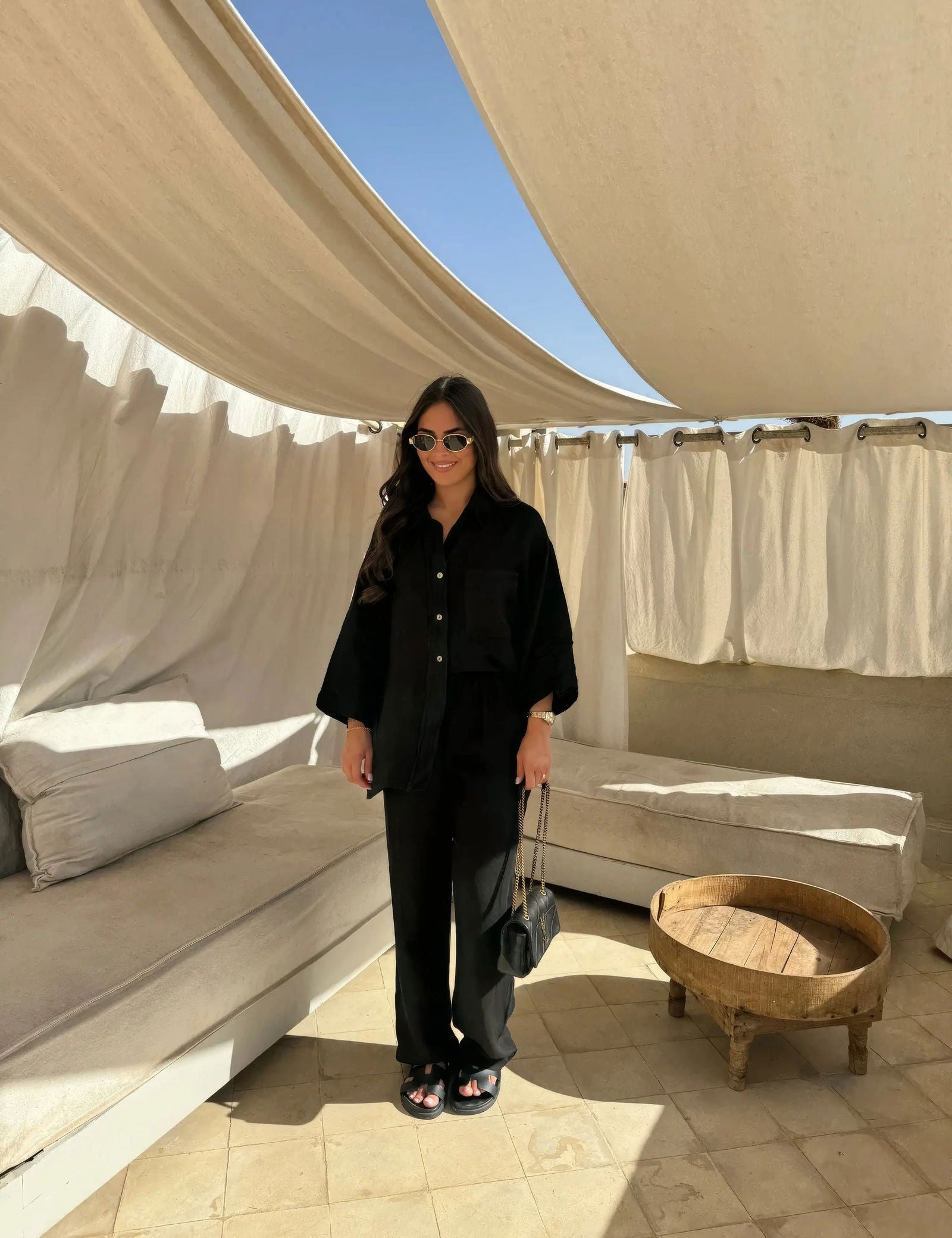 Donna con occhiali da sole indossa abito nero elegante, in terrazza con tende bianche e divani beige. Moda estiva, stile casual chic.