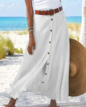 Gonna lunga bianca con bottoni verdi, cintura marrone, indossata su spiaggia tropicale. Moda estiva donna, abbigliamento casual elegante.