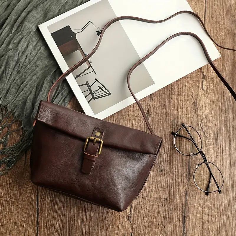 Borsa a tracolla in pelle marrone su tavolo di legno, accanto a occhiali e rivista. Accessorio moda elegante, perfetto per look casual e chic.