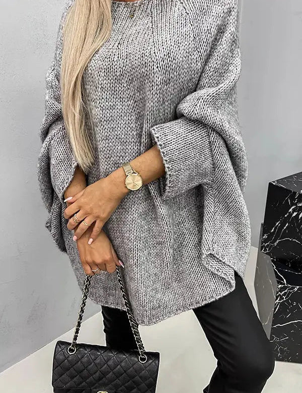 Donna con maglione oversize grigio, pantaloni neri, borsa a tracolla nera trapuntata, orologio dorato. Moda autunnale elegante e casual.