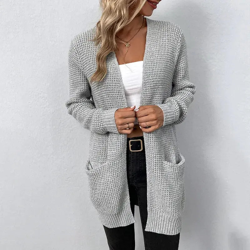 Donna con cardigan grigio lavorato a maglia, tasche frontali, indossa top bianco e jeans neri. Moda casual autunnale, abbigliamento femminile elegante.