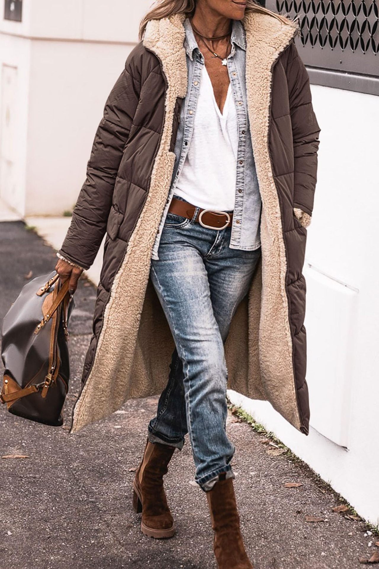 Donna con cappotto lungo marrone imbottito, jeans, stivali marroni e borsa in pelle. Moda invernale casual, abbigliamento caldo e confortevole.