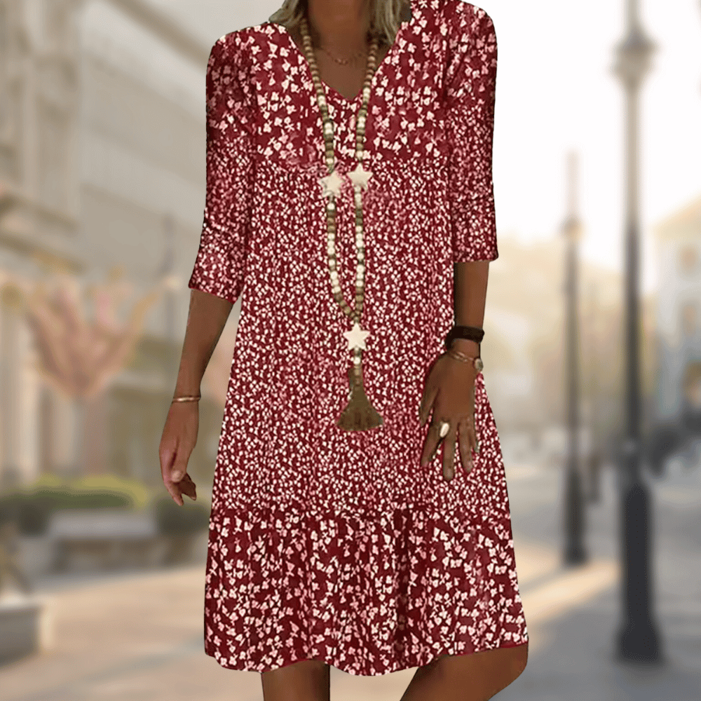 Abito estivo donna rosso a fiori, maniche a tre quarti, scollo a V, collana lunga, sfondo urbano sfocato. Moda casual elegante.