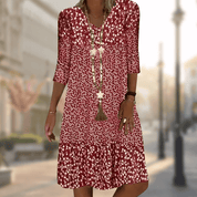 Abito estivo donna rosso a fiori, maniche a tre quarti, scollo a V, collana lunga, sfondo urbano sfocato. Moda casual elegante.