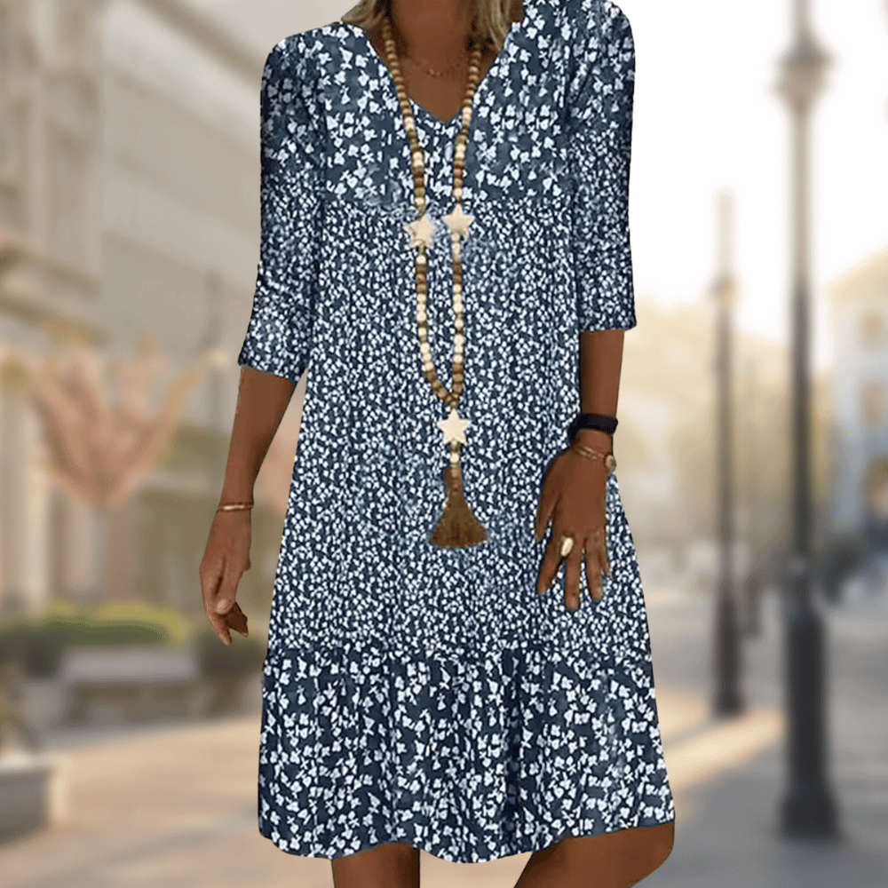 Abito estivo donna blu a fiori bianchi, maniche a tre quarti, collana lunga, sfondo urbano sfocato. Moda casual elegante.