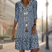 Abito estivo donna blu a fiori bianchi, maniche a tre quarti, collana lunga, sfondo urbano sfocato. Moda casual elegante.