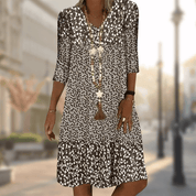 Abito estivo donna a fiori bianchi e neri, maniche a tre quarti, collana lunga, sfondo urbano sfocato. Moda casual elegante.