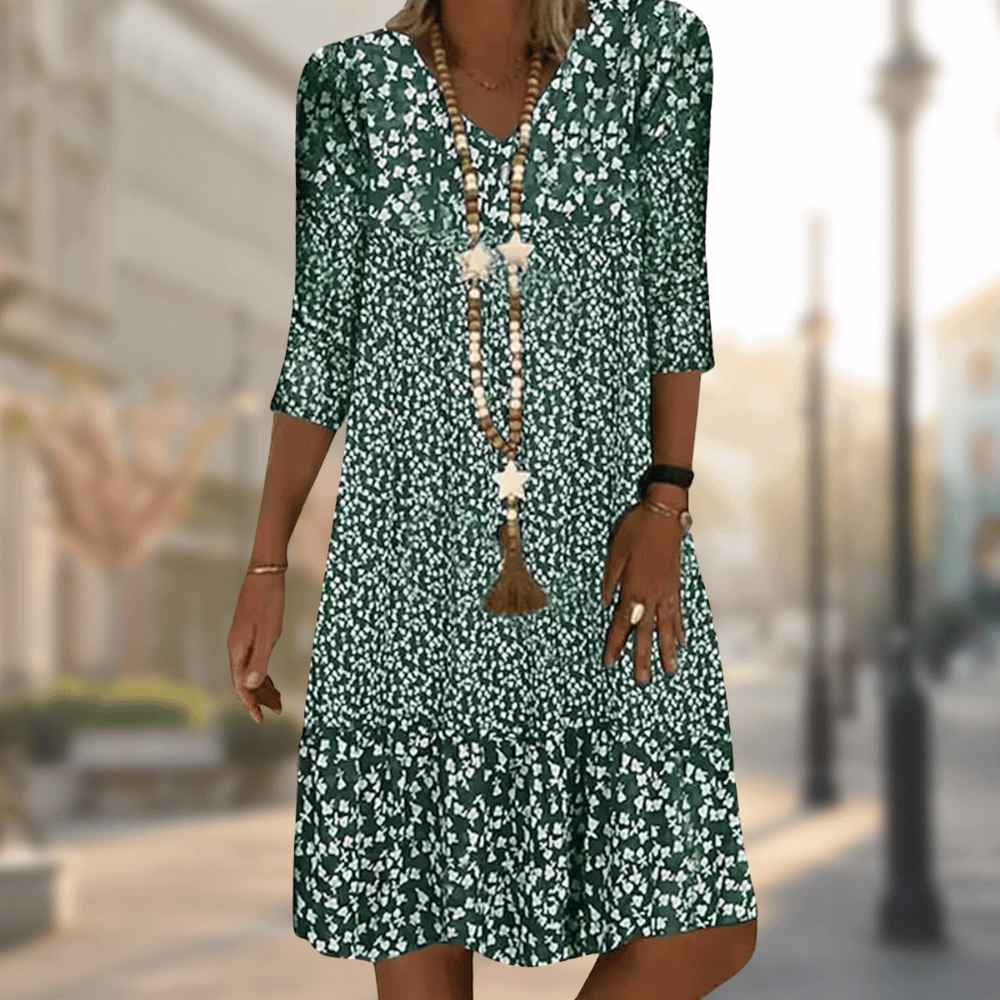 Abito estivo verde a fiori bianchi, maniche a tre quarti, scollo a V, collana lunga, sfondo urbano sfocato. Moda donna casual elegante.