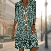 Abito estivo verde a fiori bianchi, maniche a tre quarti, scollo a V, collana lunga, sfondo urbano sfocato. Moda donna casual elegante.