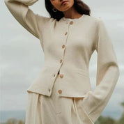Donna con cardigan beige in lana, bottoni in legno, maniche lunghe, abbinato a pantaloni coordinati. Moda autunnale elegante e minimalista.