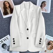 Giacca bianca elegante da donna con bottoni neri su sfondo grigio, circondata da riviste di moda. Stile moderno, ideale per look professionali.