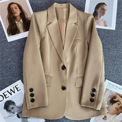 Blazer beige elegante con bottoni neri su sfondo grigio, circondato da riviste di moda. Ideale per look professionali e casual chic.