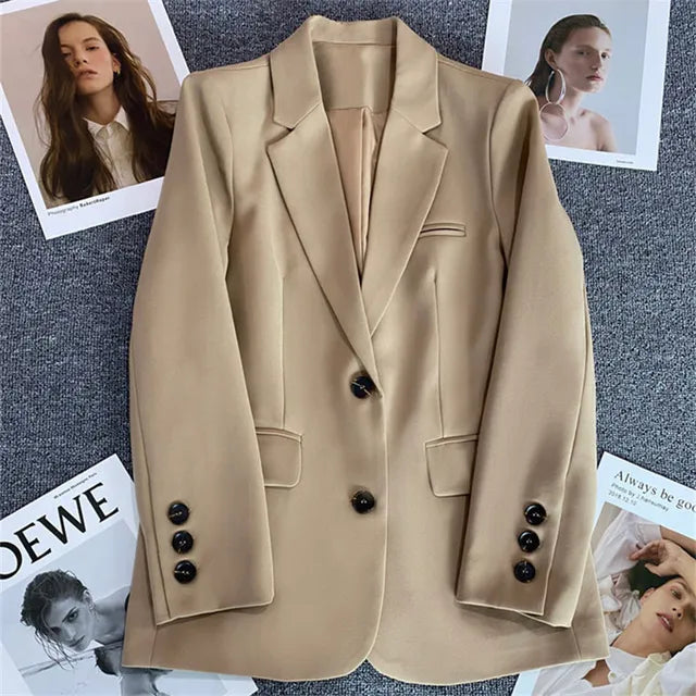 Blazer beige elegante con bottoni neri su sfondo grigio, circondato da riviste di moda. Ideale per look professionali e casual chic.