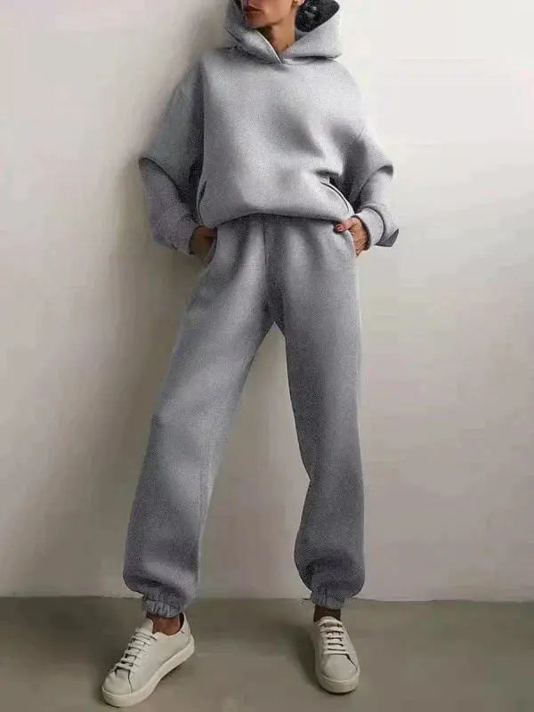 Donna in tuta sportiva grigia con cappuccio, pantaloni larghi e sneakers bianche. Abbigliamento casual, moda autunnale, stile confortevole.