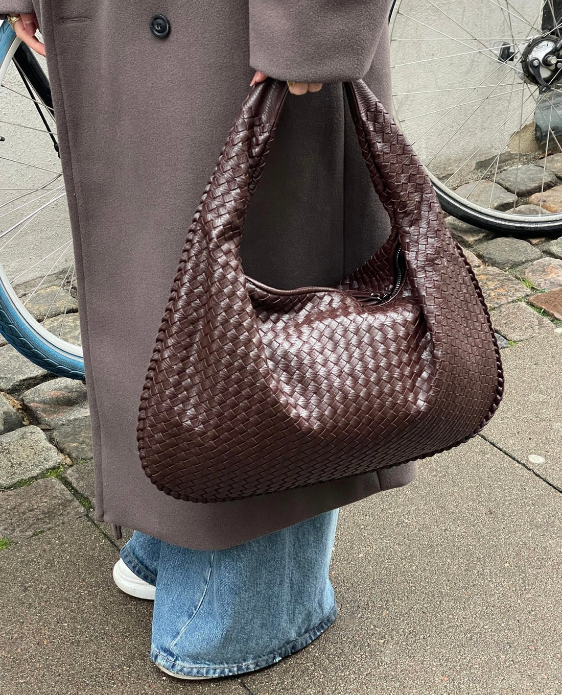 Borsa intrecciata marrone scuro in pelle, indossata da una persona con cappotto grigio e jeans, accanto a una bicicletta su marciapiede urbano.