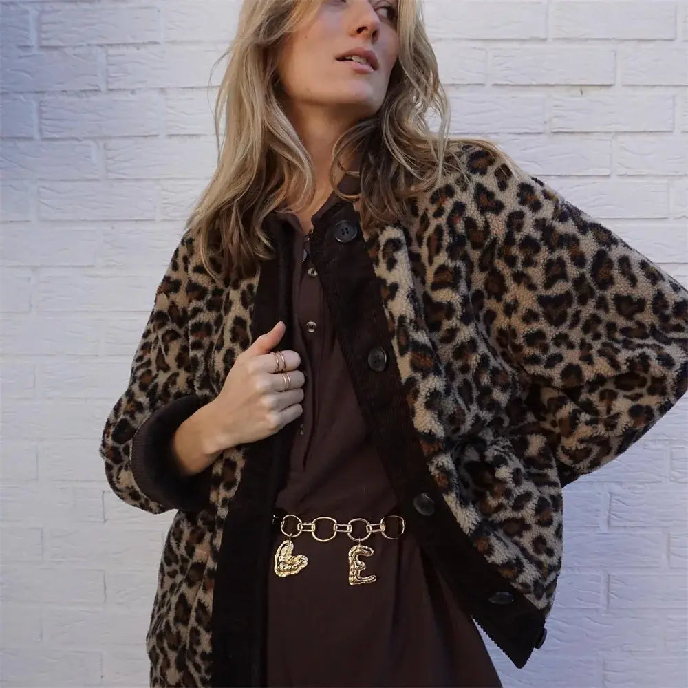 Donna con giacca leopardata e cintura dorata su abito marrone, posa davanti a muro bianco. Moda autunno, stile casual chic, tendenze 2023.