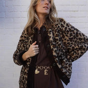Donna con giacca leopardata e cintura dorata su abito marrone, posa davanti a muro bianco. Moda autunno, stile casual chic, tendenze 2023.