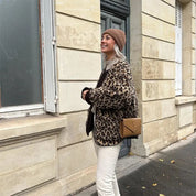 Donna con cappotto leopardato, berretto marrone e borsa a tracolla beige, cammina su marciapiede urbano. Moda autunnale, stile casual chic.