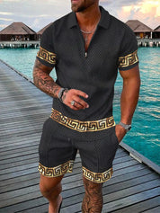 Uomo in completo estivo nero con motivi dorati, camicia a polo e pantaloncini coordinati, su passerella di legno vicino al mare. Moda estiva elegante.
