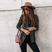 Donna alla moda con cappello marrone, occhiali da sole, giacca a quadri verde e borsa a tracolla rosa, appoggiata a un muro di cemento.
