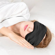 Donna che dorme con maschera per dormire nera in seta, indossa pigiama bianco, su letto bianco. Accessorio relax, miglioramento sonno, comfort notturno.