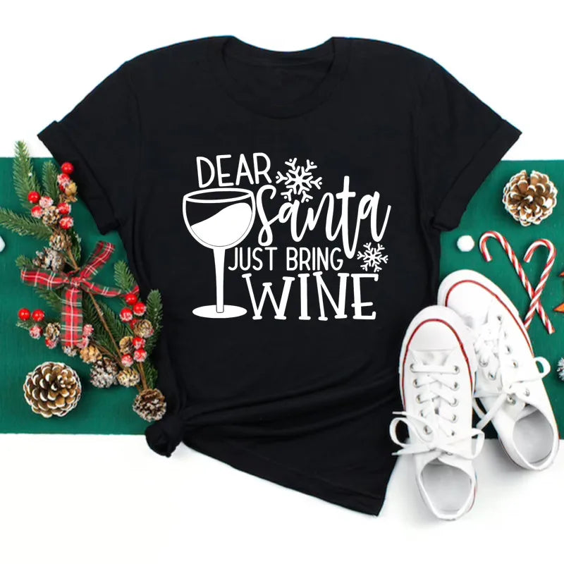 Maglietta nera con scritta "Dear Santa Just Bring Wine", decorata con bicchiere di vino e fiocchi di neve, accanto a scarpe bianche e decorazioni natalizie.