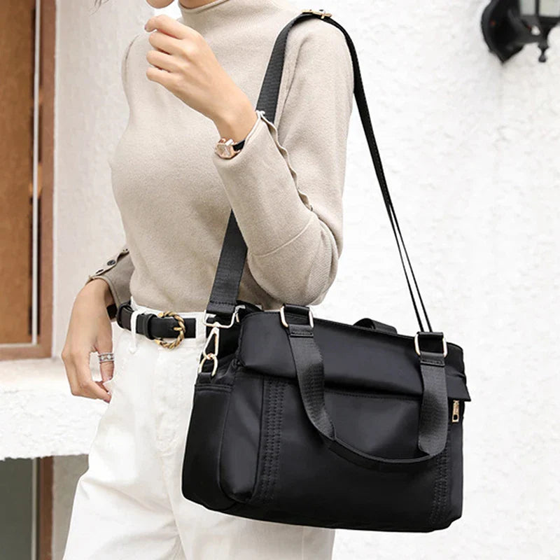 Donna con borsa a tracolla nera in tessuto, indossa maglione beige e pantaloni bianchi. Accessorio moda elegante e pratico per uso quotidiano.