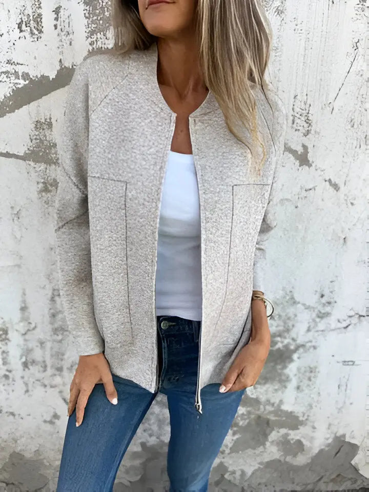 Donna con giacca beige aperta su top bianco e jeans blu, posa casual davanti a muro grigio. Moda autunnale, abbigliamento casual chic.