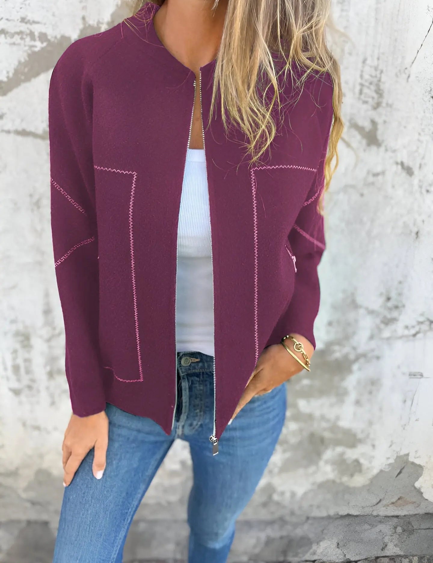 Donna con cardigan bordeaux aperto, top bianco e jeans blu. Moda casual autunnale, abbigliamento femminile elegante. Sfondo muro grigio.