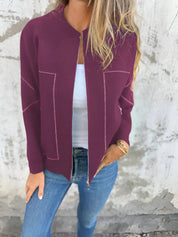 Donna con cardigan bordeaux aperto, top bianco e jeans blu. Moda casual autunnale, abbigliamento femminile elegante. Sfondo muro grigio.