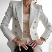 Giacca bianca elegante con bottoni dorati, indossata con top coordinato. Accessori oro e anello con pietra rosa. Moda donna, stile sofisticato.