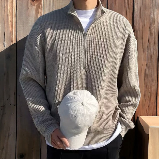 Uomo con maglione beige a collo alto e zip, tiene un cappello bianco. Sfondo in legno. Moda casual autunnale, abbigliamento maschile elegante.