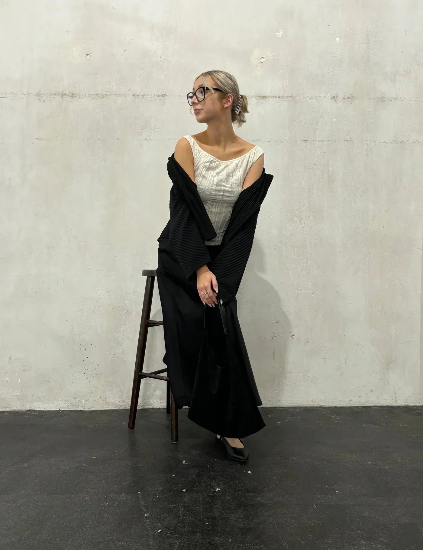 Donna elegante con occhiali, abito bianco e nero, appoggiata a uno sgabello su sfondo neutro. Moda contemporanea, stile minimalista.