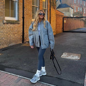 Donna con occhiali da sole indossa giacca grigia oversize, leggings blu scuro e scarpe da ginnastica grigie, cammina su strada asfaltata.