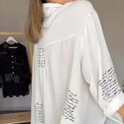 Camicia bianca con scritte nere, maniche lunghe, design elegante. Sullo sfondo, camicia nera appesa. Moda femminile, abbigliamento casual.