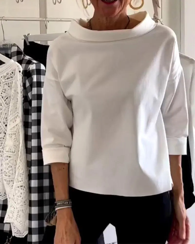 Donna indossa maglia bianca a maniche lunghe con collo alto, abbinata a pantaloni neri. Sfondo con abiti a quadri e pizzo. Moda casual elegante.