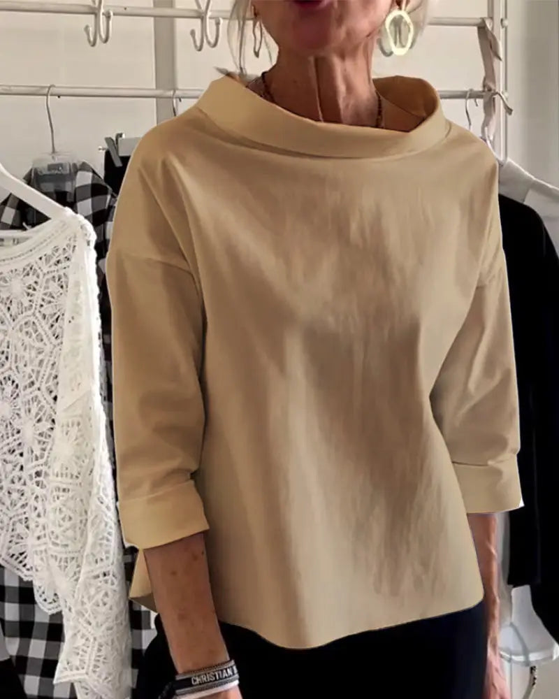 Donna indossa maglia beige a maniche lunghe, collo ampio, su sfondo con abiti appesi. Moda femminile, abbigliamento casual elegante.