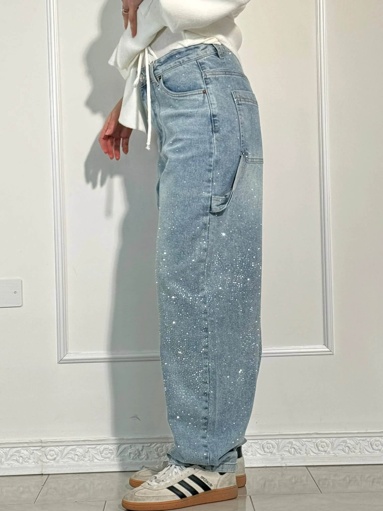 Jeans a vita alta in denim chiaro con dettagli glitterati, tasche laterali, indossati con sneakers bianche e nere, sfondo bianco minimalista.